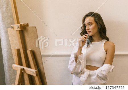 Young female artist standing near easel and holding pencil while working on painting in art studio, creative process and inspiration Young female artist standing near easel and holding pencil while working on painting in art studio, creative process and inspiration 137347723