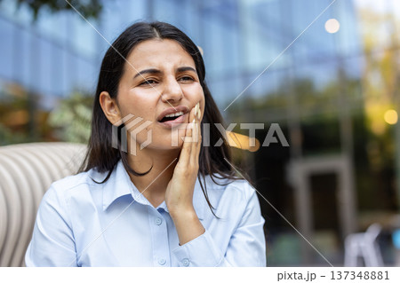 Young woman experiencing sharp pain from a toothache, grimacing and touching her jaw with her hand, highlighting dental issues and oral health problems outdoors 137348881