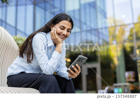 Businesswoman sitting outside a modern office building. Smiling and laughing while using her smartphone and wireless earbuds. Conveying connectivity. Digital communication. And relaxed professionalism Businesswoman sitting outside a modern office building. Smiling and laughing while using her smartphone and wireless earbuds. Conveying connectivity. Digital communication. And relaxed professionalism 137348887