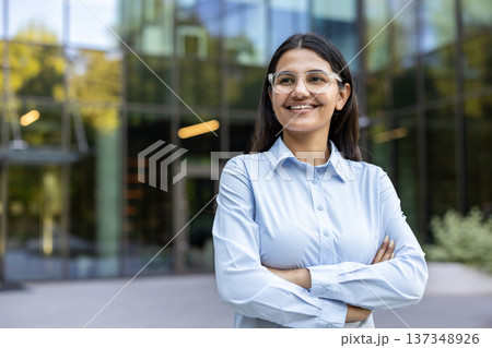 Young indian businesswoman in a light blue shirt and clear glasses smiling confidently with arms crossed, standing in front of a modern glass office building Young indian businesswoman in a light blue shirt and clear glasses smiling confidently with arms crossed, standing in front of a modern glass office building 137348926