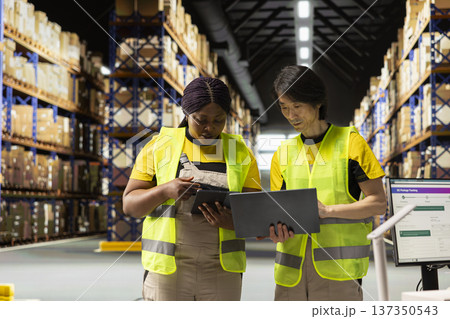 Multiethnic staff processing e-commerce orders and reviewing invoices, organizing cargo goods inside industrial warehouse space filled with storage racks. Smooth delivery and quality control. 137350543