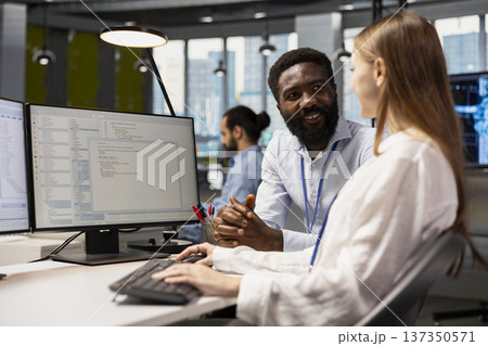 Happy senior programmer mentoring junior developer at desk in office, teaching best coding practices. Cheerful tech company lead engineer guiding intern through project tasks 137350571