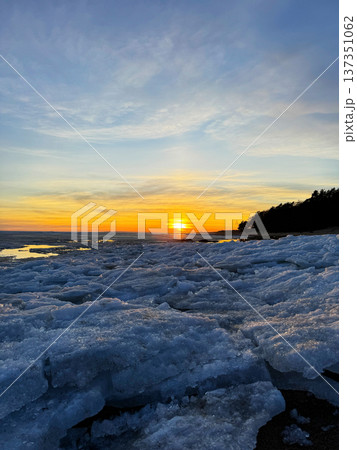 Beautiful winter sunset over a frozen body of water, likely large lake or bay. The icy foreground contrasts sharply with the golden light of the setting sun and the dark shoreline treeline. 137351062