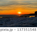 Serene winter sunset over frozen body of water, likely a lake or sea, with large chunks of blue and white ice covering foreground. Silhouetted trees line the distant shore, creating natural contrast. 137351548