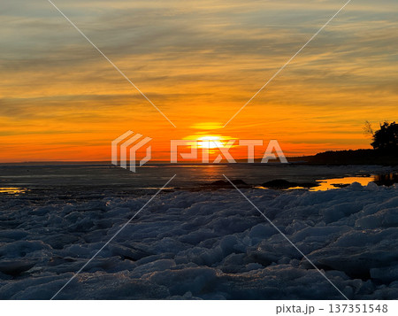 Serene winter sunset over frozen body of water, likely a lake or sea, with large chunks of blue and white ice covering foreground. Silhouetted trees line the distant shore, creating natural contrast. Serene winter sunset over frozen body of water, likely a lake or sea, with large chunks of blue and white ice covering foreground. Silhouetted trees line the distant shore, creating natural contrast. 137351548