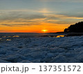 Vibrant winter sunset casts warm orange and yellow light across partially frozen body of water, with large chunks of ice and dark, tree-lined shoreline silhouetted in the distance.  137351572