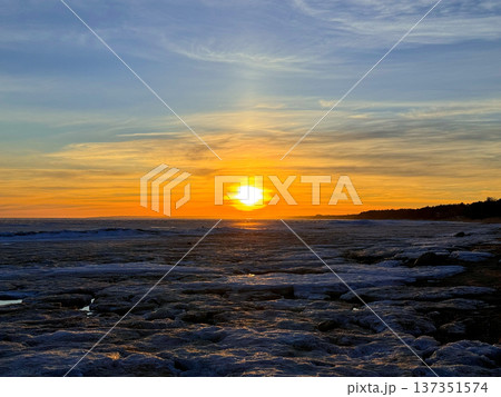 Vibrant sunset illuminates icy shore, painting the sky with vivid orange and yellow hues. The sun sets over frozen pond. Scene is cold and beautiful. Winter landscape.  137351574