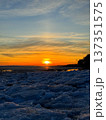 Fiery winter sunset over frozen body of water, likely a lake or sea, with large chunks of blue and white ice covering foreground. Silhouetted trees line the distant shore, creating natural contrast. 137351575