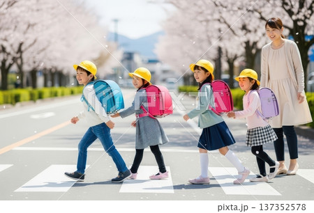 桜の花びらが舞う春の通学路で黄色い帽子とランドセルの新一年生たちと先生や保護者 137352578