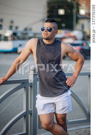 Portrait of a muscular man wearing a tank top and shorts, leaning on a railing on a sunny day in a russian city 137356388