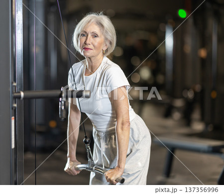In gym, senior woman uses cable row machine to strengthen muscles. 137356996