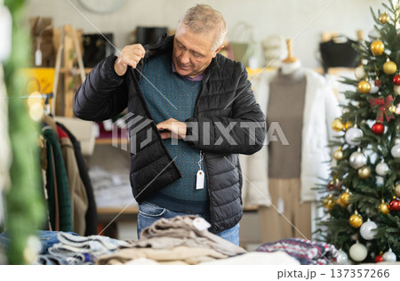 Elderly man chooses warm down jacket in clothing store. Elderly man chooses warm down jacket in clothing store. 137357266