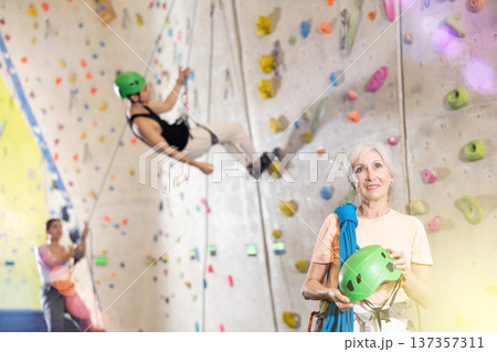 Smiling active mature woman standing with rope on shoulders and helmet in hand against artificial training climbing wall in adventure park 137357311