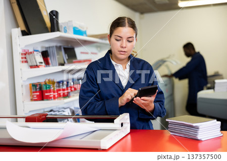 Focused young female typographer in uniform calculates the cost of printed notepads and sheet in the typography 137357500
