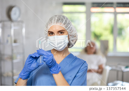 Nurse in blue uniform with mask and gloves behind hat holding syringe with medicine, patient behind. Nurse in blue uniform with mask and gloves behind hat holding syringe with medicine, patient behind. 137357544