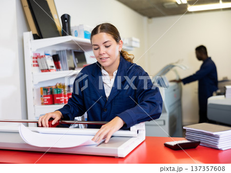 Portrait of cheerful young female typographer in uniform using paper cutter during work in printer shop Portrait of cheerful young female typographer in uniform using paper cutter during work in printer shop 137357608