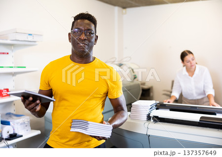 Happy middle-aged African American male typographer showing new notepads in the middle of the copy center Happy middle-aged African American male typographer showing new notepads in the middle of the copy center 137357649