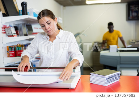 Positive female employee at printing house cuts paper with paper cutter Positive female employee at printing house cuts paper with paper cutter 137357682