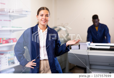 Cheerful young woman in blue robe uniform sticking out her left hand to show the workflow in the printer house Cheerful young woman in blue robe uniform sticking out her left hand to show the workflow in the printer house 137357699