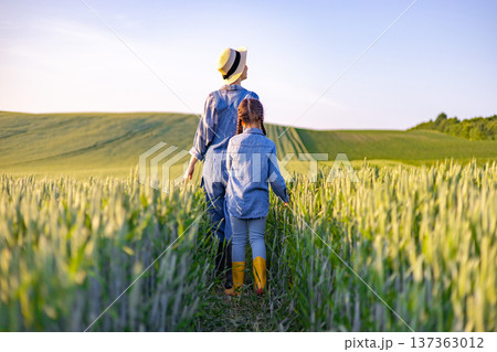 A mother and her daughter walk through a golden wheat field under a clear blue sky, enjoying a serene rural landscape 137363012