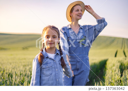 A mother and daughter stand in a sunlit field of green crops, both dressed in denim overalls and shirts, with the daughter in the foreground and the mother behind her wearing a straw hat A mother and daughter stand in a sunlit field of green crops, both dressed in denim overalls and shirts, with the daughter in the foreground and the mother behind her wearing a straw hat 137363042