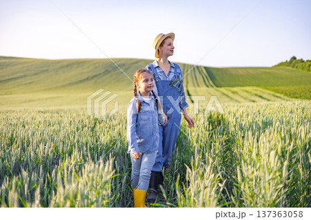 A woman and a young girl stand in a vast field of wheat, looking towards the horizon under a clear sky A woman and a young girl stand in a vast field of wheat, looking towards the horizon under a clear sky 137363058