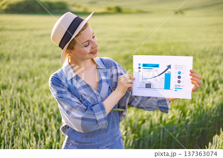 A woman in a straw hat and overalls stands in a field of green crops, holding up a paper with a rising bar graph and statistics A woman in a straw hat and overalls stands in a field of green crops, holding up a paper with a rising bar graph and statistics 137363074