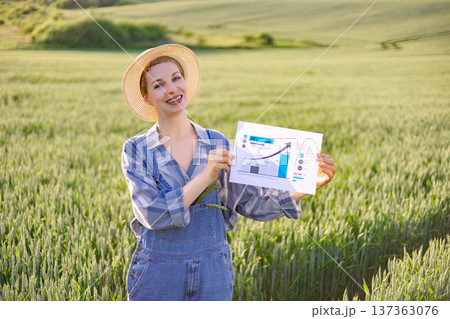 A smiling woman in overalls and a straw hat stands in a green field, holding a paper with a growing bar chart and arrow 137363076