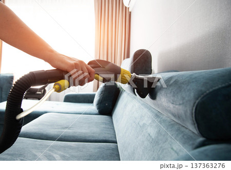 Hand of person using wet vacuum cleaner tool for deep cleaning of upholstered blue sofa in living room during day 137363276