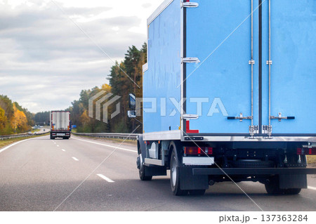 Blue delivery truck rear view driving on multi-lane highway during day with autumn color trees lining roadside under overcast sky freight transportation logistics commerce distribution service 137363284