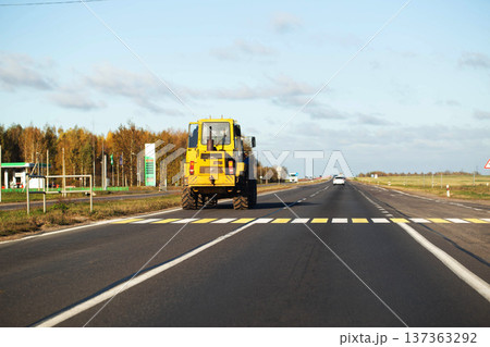 Yellow tractor driving on highway road during daylight hours with gas station and autumn trees in background, other vehicles in distance, wide view of rural landscape. 137363292