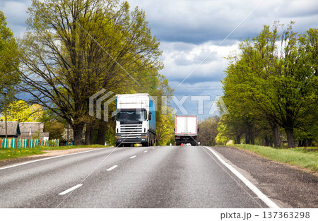 Long haul trucks traveling rural road lined with lush green trees and traditional houses under cloudy spring sky. Heavy freight transport. Long haul trucks traveling rural road lined with lush green trees and traditional houses under cloudy spring sky. Heavy freight transport. 137363298
