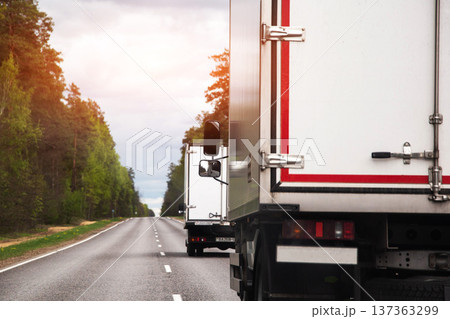White cargo trucks traveling down rural highway, tree line alongside asphalt road, cloudy sky above, logistics and freight industry concept. White cargo trucks traveling down rural highway, tree line alongside asphalt road, cloudy sky above, logistics and freight industry concept. 137363299