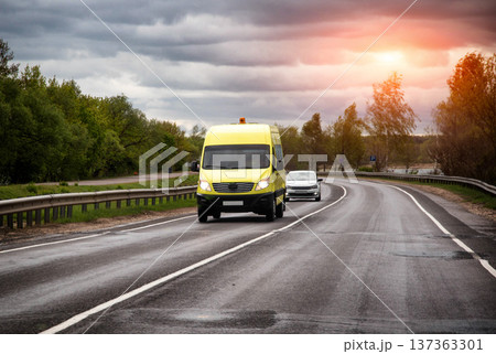 Yellow school bus drives wet asphalt road cloudy sky, white passenger car follows closely behind, green trees line winding rural roadway. 137363301