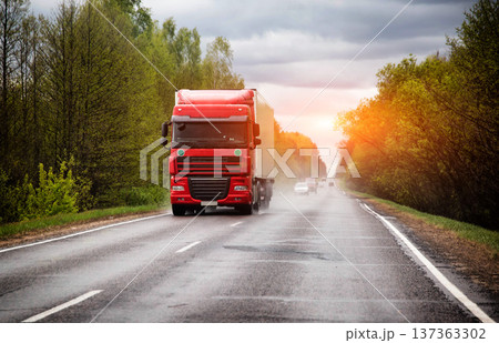Red semi-trailer truck on wet highway, green trees line road, sun light at horizon, transport and logistics business. Red semi-trailer truck on wet highway, green trees line road, sun light at horizon, transport and logistics business. 137363302