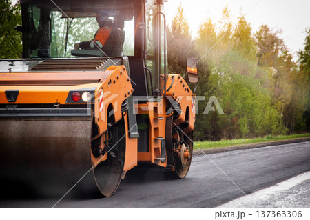 Orange road roller compacting fresh black asphalt during highway construction project. Sunlight flares from trees creating warm light. 137363306