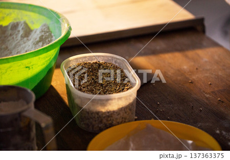 Kitchen scene shows preparation of ingredients for baking or cooking process, display of flour in vibrant green bowl, diverse seeds in clear plastic container on rustic wooden table. 137363375