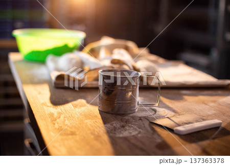 Metal flour sifter on wooden surface with scattered flour, dough portions, and green bowl in soft focus background, highlighting preparation process of baking bread or pastry 137363378