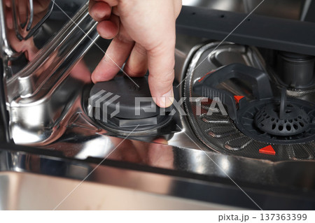 Close-up shot of human hand unscrewing cap of salt dispenser in bottom part of modern dishwasher for refilling appliance with special cleaning agent. 137363399