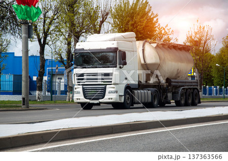 White semi-truck with tanker trailer travels on multi-lane road during daytime. Trees line side of road. Sun flare visible. Flags on pole. White semi-truck with tanker trailer travels on multi-lane road during daytime. Trees line side of road. Sun flare visible. Flags on pole. 137363566