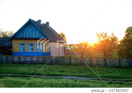Rural house with vibrant blue and yellow facade and old wooden fence at sunset. Green field of dandelions in foreground. Peaceful countryside landscape during golden hour. Rural house with vibrant blue and yellow facade and old wooden fence at sunset. Green field of dandelions in foreground. Peaceful countryside landscape during golden hour. 137363567