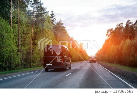 Black cargo van driving on rural road toward horizon light of sunset. Other truck in distance. Lush green forest line road journey freight logistics Black cargo van driving on rural road toward horizon light of sunset. Other truck in distance. Lush green forest line road journey freight logistics 137363568
