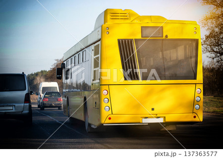 Bright yellow public transport bus moving on asphalt road, showing rear of vehicle with taillights, other automobiles following closely in traffic, outdoor daytime setting Bright yellow public transport bus moving on asphalt road, showing rear of vehicle with taillights, other automobiles following closely in traffic, outdoor daytime setting 137363577