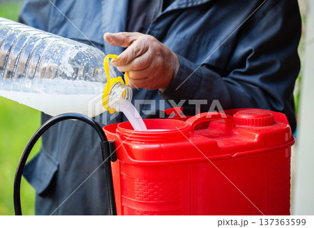 Person hand pouring white solution from plastic bottle into red garden sprayer, preparing for thorough plant protection activity. 137363599