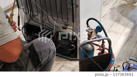 Refrigerator repair professional at work, domestic appliance back panel removed, showing compressor, condenser coils, using refrigerant tanks, pressure gauges, hoses, welding torch for service 137363691