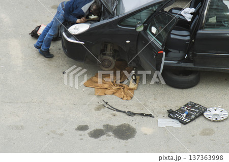 Man repairing engine of black car outdoors, front wheel removed, car lifted on jack for maintenance. Tools and various car parts scattered on asphalt ground, oil stains visible near vehicle. 137363998
