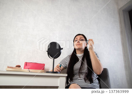 Adult woman carefully applies mascara to upper eyelashes looking into round tabletop mirror, white dressing table with cosmetic bag and brush, indoor beauty routine. Copy space for text 137363999