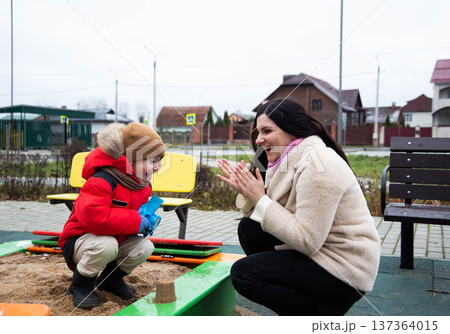 Young boy in red winter coat with adult woman in beige coat laughing and clapping hands at sandbox playtime during overcast day outdoors 137364015