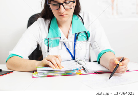 Female medical professional focused on paperwork, reviewing patient records, making notes, performing essential medical office tasks. 137364023