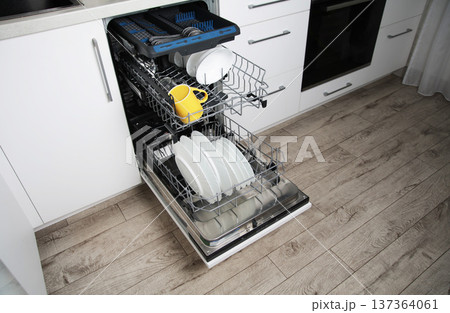 Open modern built-in dishwasher in white kitchen interior with white plates yellow mug cutlery on racks ready for washing cycle startup 137364061